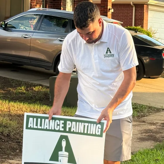 An Alliance Painting team member in a branded polo planting the company yard sign on a front lawn in evening light