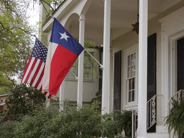 A Texas flag and a United States flag on the porch of a white-painted brick home