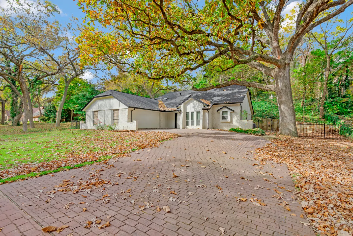 Single-story home under mature oaks after repainting, with a brick paver driveway and autumn leaves across the lawn