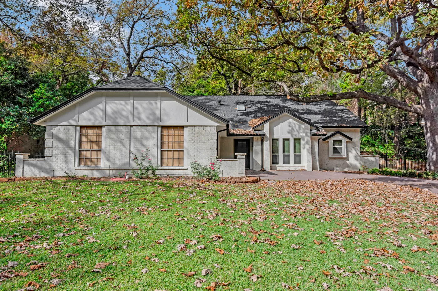 Front of a repainted brick ranch house with a board-and-batten gable and tall windows, framed by oak branches