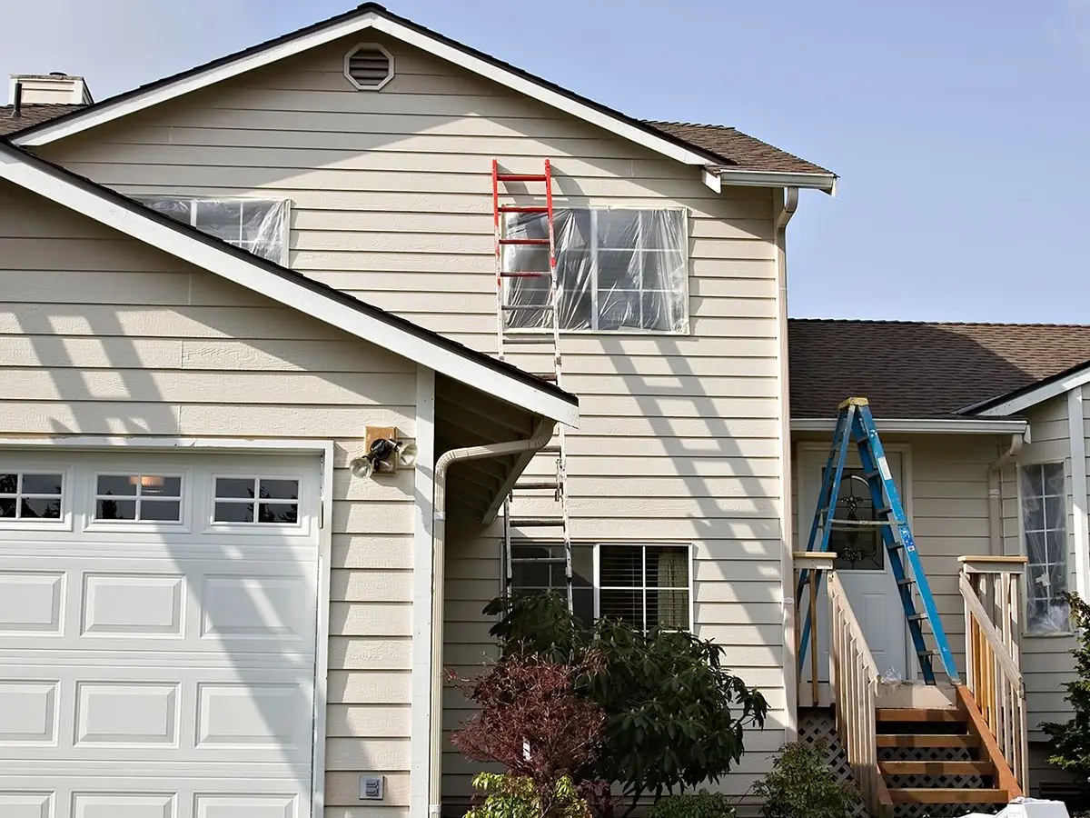 Residential house / home being prepared for exterior painting with windows covered / masked with plastic and several ladders against siding