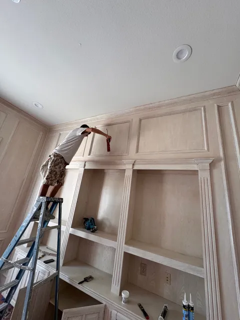 A painter on a step ladder brushing the upper rail of a floor-to-ceiling built-in bookcase