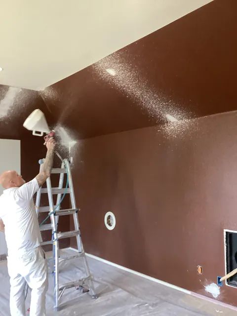A painter spraying the ceiling of a room with deep brown walls, the spray mist caught in the light