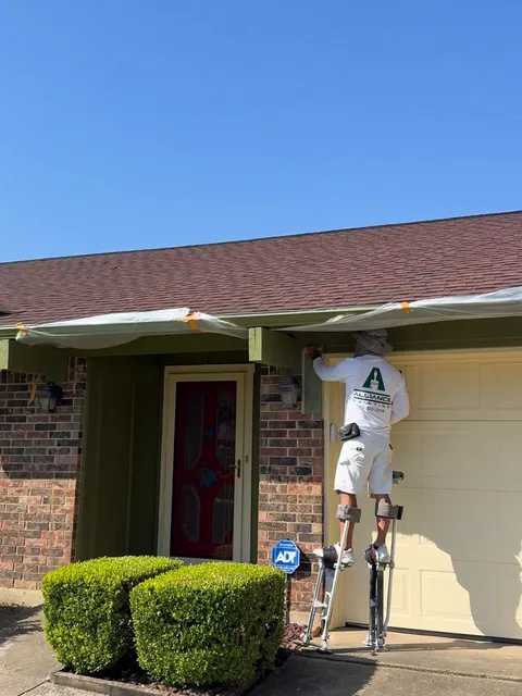 A single-story brick house part-way through an exterior repaint, a painter on stilts cutting in the beam beneath a masked roofline