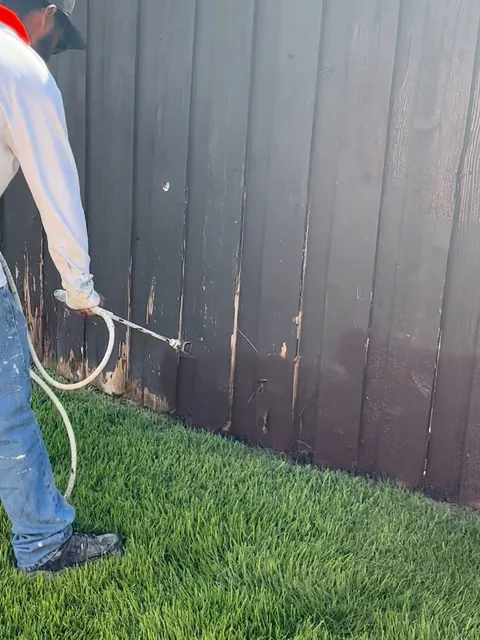 A spray gun laying stain onto a board fence, with the dry weathered boards still visible below the wet line