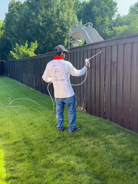 A long run of board fence being stained, seen down its length in full sun
