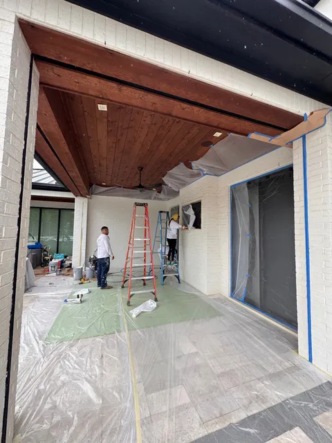 A covered patio with a stained tongue-and-groove ceiling and painted brick walls, two crew members working at the far end