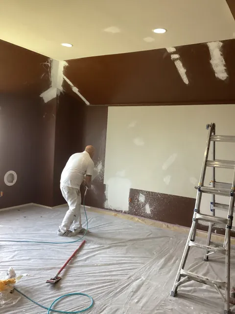 A painter rolling a deep brown wall in an empty room, the patched drywall above it still showing filler